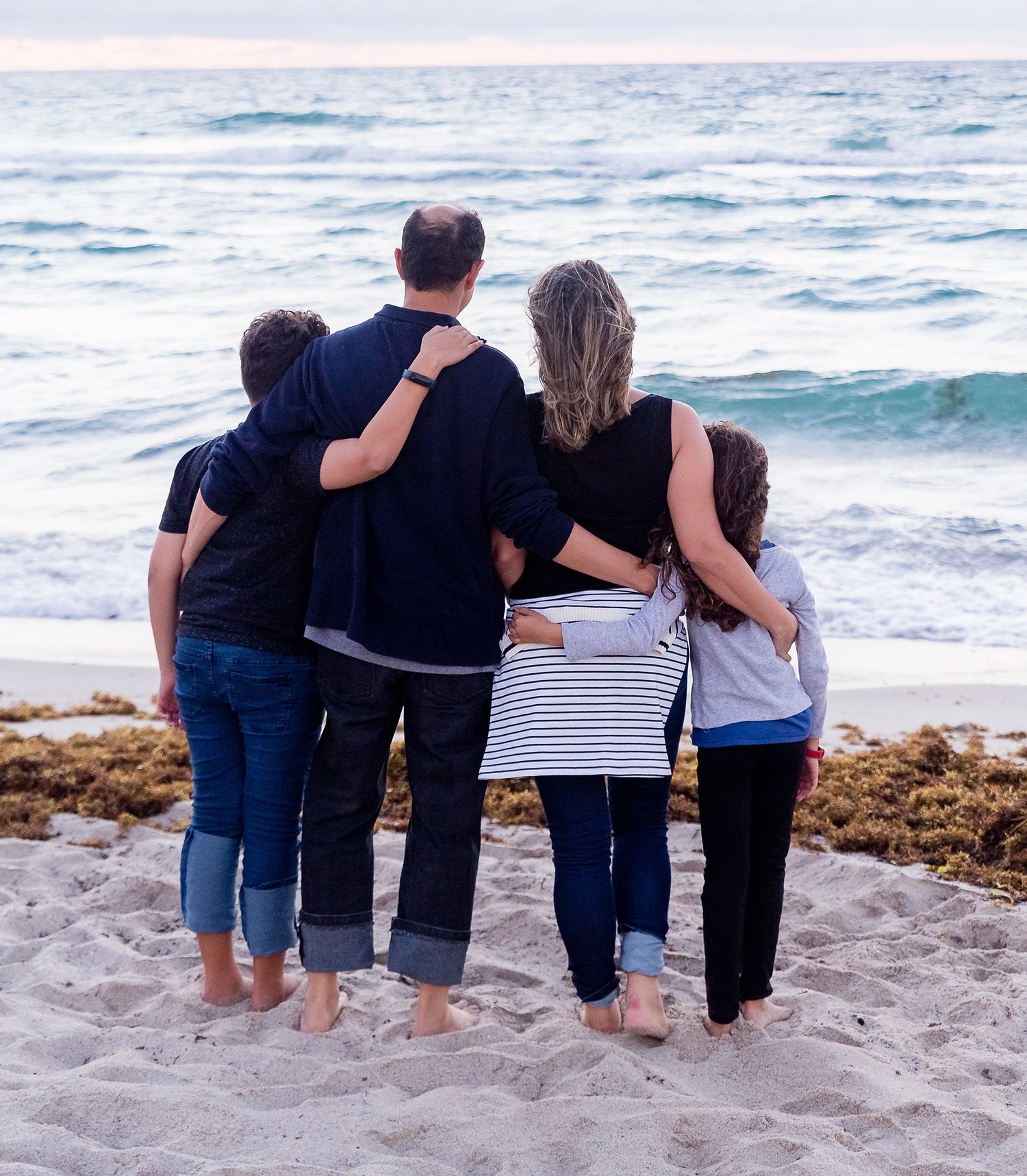 Frau mit Mann und zwei Kindern Arm in Arm am Strand blicken aufs Meer